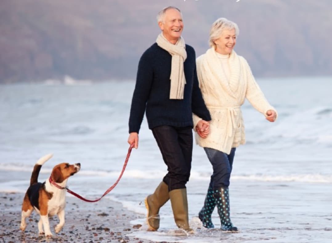 Two older people, man and woman, walking hand-in-hand on a beach. The man holds the leash of a dog and has a very large smile. The woman appears to be wearing a bathrobe.