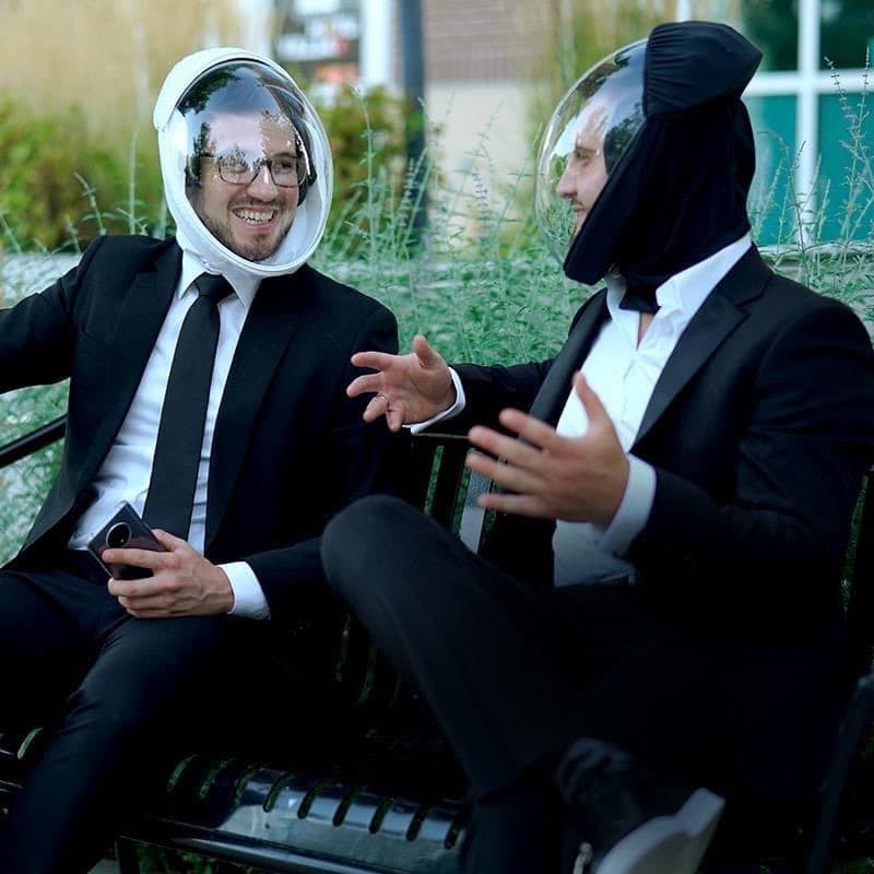 Two tech bros conversing on a bench. They both wear suits, and ridiculous helments with clear shields covering their faces. Covid-19 relic.